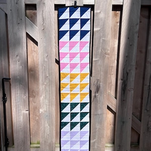 May include: A colorful, geometric quilt hanging on a wooden fence. The quilt features alternating rows of triangles in navy blue, pink, yellow, green, and lavender, with white polka dots. The quilt is bordered in black.