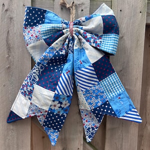 May include: A large, handmade bow made of various blue and white patterned fabrics, including polka dots, floral prints, and stripes. The bow is attached with a twine loop, perfect for wall decor or seasonal accents.