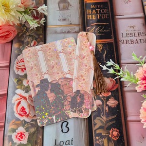 May include: Three romantic bookmarks with a couple silhouette design and glitter accents. They have a floral and striped pattern, and a tassel with pink beads. The background includes vintage books and floral arrangements.