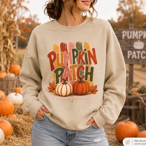 Könnte beinhalten: Ein beigefarbenes Sweatshirt mit Rundhalsausschnitt und einem Grafikdruck mit dem Text "PUMPKIN PATCH" in gestapelten, bunten, abgenutzten Buchstaben. Unter dem Text befinden sich zwei Kürbisse, einer weiß und einer orange, umgeben von Herbstblättern und dem Text "AUTUMN HARVEST".