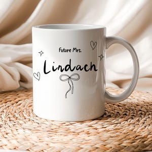 Könnte beinhalten: Ein weißer Keramikbecher mit schwarzem Aufdruck Future Mrs. Lindach. Das Design umfasst eine zentrale schwarze Strichzeichnung einer Schleife, zwei kleine Herzumrisse und zwei vierzackige Sternsymbole, die um den Schriftzug angeordnet sind.