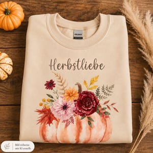 Könnte beinhalten: Ein beigefarbenes Sweatshirt mit Rundhalsausschnitt und dem Schriftzug "Herbstliebe" in dunkelbrauner Schreibschrift. Unter dem Text befindet sich eine Aquarell-Illustration eines großen orangefarbenen Kürbisses mit herbstlichen Blumen.
