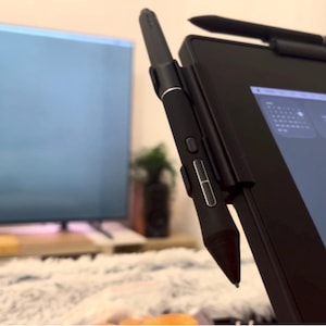 May include: A black digital stylus with two silver-accented side buttons attached to the side of a black tablet screen. The stylus has a tapered tip and a cylindrical body, positioned vertically against the edge of the dark electronic display device.