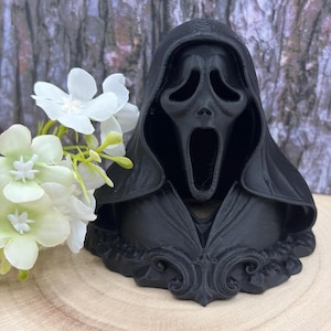 May include: A black Ghostface mask bust from the Scream film series. The mask features a gaping mouth and empty eye sockets. The bust is displayed on a wooden surface, accompanied by white flowers.