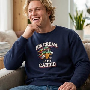 Op de afbeelding: Een marineblauwe sweater met ronde hals voorzien van een afbeelding van een rennend ijsje met een zonnebril en een hoofdband, met de tekst "ICE CREAM IS MY CARDIO" in witte en rode letters op de borst van het kledingstuk gedrukt.