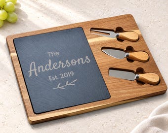 May include: A rectangular wooden serving board featuring a dark gray slate inlay engraved with the text The Andersons Est. 2019 and a leaf graphic. The board includes three recessed slots containing a cheese fork and two cheese knives.