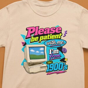 以下が含まれることがあります： アイボリーのTシャツで、ベージュのヴィンテージコンピューターのモニターとタワーのグラフィックプリントが施されています。テキストには「Please be patient with me. I'm from the 1900's」と書かれています。デザインには、明るいピンク、青、黄色、紫のレトロなアクセントが含まれています。