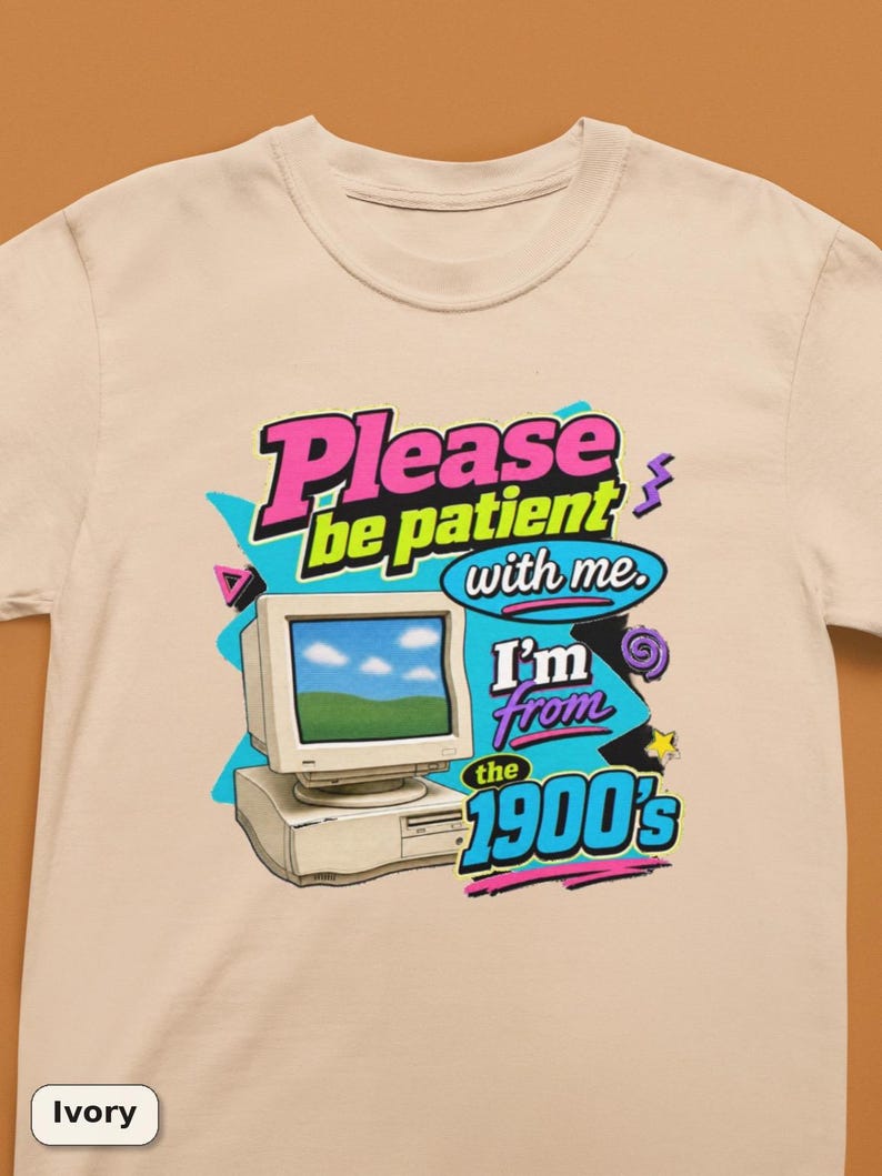 以下が含まれることがあります： アイボリーのTシャツで、ベージュのヴィンテージコンピューターのモニターとタワーのグラフィックプリントが施されています。テキストには「Please be patient with me. I'm from the 1900's」と書かれています。デザインには、明るいピンク、青、黄色、紫のレトロなアクセントが含まれています。