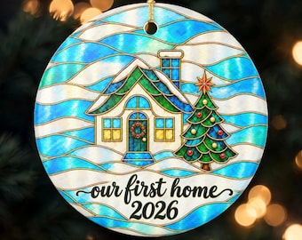May include: A round ceramic ornament featuring a stained-glass style illustration of a house and a decorated Christmas tree. The background has blue and white wavy lines. The text reads our first home 2026 in black script and print.
