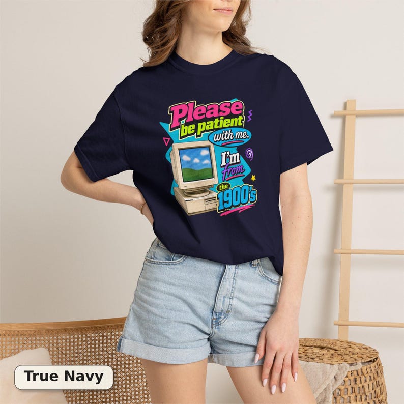 以下が含まれることがあります： ネイビーブルーの半袖Tシャツ。風景を表示したベージュのヴィンテージコンピューターモニターのグラフィックが特徴です。テキストは「Please be patient with me. I'm from the 1900's」と書かれており、ピンク、青、紫の幾何学模様がアクセントになっています。