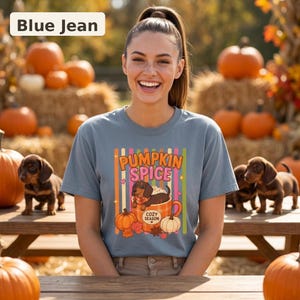 May include: A blue jean colored t-shirt featuring a graphic of a dachshund sitting in a mug with the text PUMPKIN SPICE and COZY SEASON. The design includes vertical stripes, pumpkins, and autumn leaves, centered on the front of the garment.