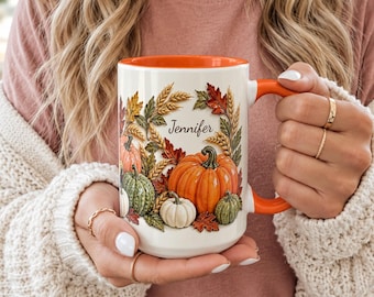 May include: A white ceramic mug with an orange interior and handle, featuring a printed design of various pumpkins, wheat stalks, and autumn leaves surrounding the name Jennifer in a black cursive script font on the front surface.