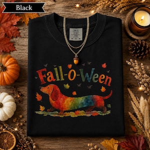 May include: A black t-shirt featuring a colorful watercolor-style dachshund dog graphic and the text "Fall-o-WEEN" in a multi-colored font. The design includes small autumn leaves scattered around the dog, with a gold-toned acorn pendant necklace.