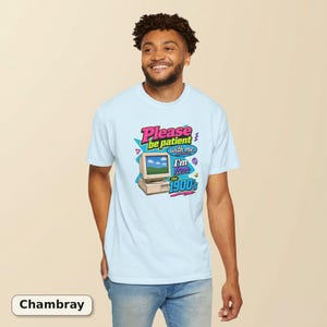 以下が含まれることがあります： 淡いブルーのTシャツ。ベージュのヴィンテージ風コンピューターモニターのグラフィックと、「Please be patient with me. I'm from the 1900's」というピンク、黄色、青の文字がプリントされており、小さな幾何学模様や渦巻きがアクセントになっています。