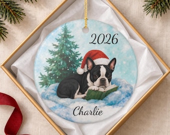 May include: A round ceramic ornament featuring a black and white French Bulldog wearing a red Santa hat, lying on snow while reading a green book. A green pine tree is in the background. The text 2026 and the name Charlie are printed on it.