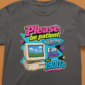 以下が含まれることがあります： チャコールグレーのTシャツで、ベージュのヴィンテージコンピューターのグラフィックが描かれています。テキストは「Please be patient with me. I'm from the 1900's.」と書かれています。デザインには、レトロなスタイルの鮮やかなピンク、イエロー、シアンのアクセントが含まれています。