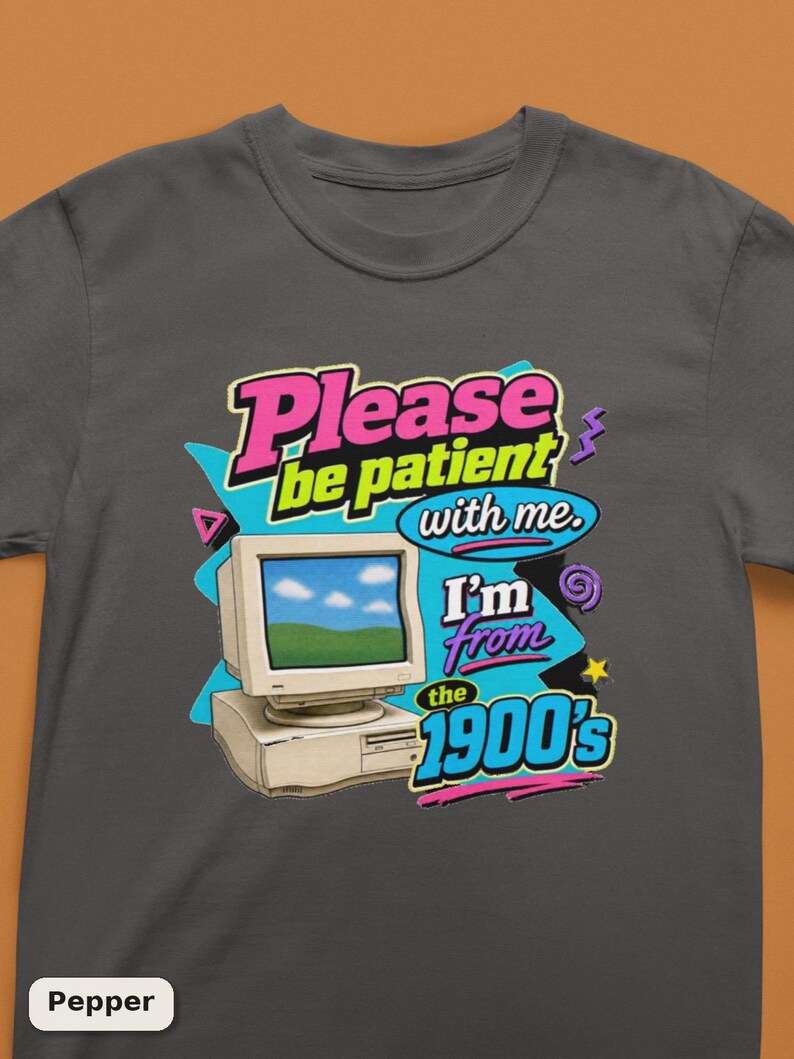 以下が含まれることがあります： チャコールグレーのTシャツで、ベージュのヴィンテージコンピューターのグラフィックが描かれています。テキストは「Please be patient with me. I'm from the 1900's.」と書かれています。デザインには、レトロなスタイルの鮮やかなピンク、イエロー、シアンのアクセントが含まれています。