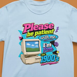 以下が含まれることがあります： ライトブルーのTシャツで、ベージュのヴィンテージコンピューターのモニターとデスクトップタワーのグラフィックが描かれています。テキストは「Please be patient with me. I'm from the 1900's」とカラフルな太字で書かれており、ピンク、紫、ターコイズの抽象的な図形で装飾されています。