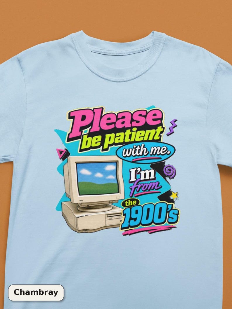 以下が含まれることがあります： ライトブルーのTシャツで、ベージュのヴィンテージコンピューターのモニターとデスクトップタワーのグラフィックが描かれています。テキストは「Please be patient with me. I'm from the 1900's」とカラフルな太字で書かれており、ピンク、紫、ターコイズの抽象的な図形で装飾されています。