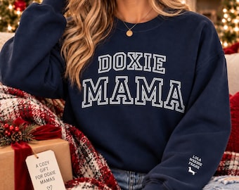 May include: A navy blue crewneck sweatshirt featuring the white text DOXIE MAMA in a collegiate font on the chest. The left sleeve includes the white text LOLA WEENIE FRANK above a small white silhouette of a dachshund dog.