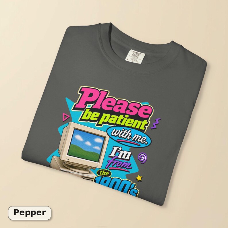 以下が含まれることがあります： チャコールグレーのTシャツ。緑の風景と青い空が映るベージュのヴィンテージ風コンピューターモニターのグラフィックが描かれています。テキストは「Please be patient with me. I'm from the 1900's.」と書かれており、ネオンピンク、ブルー、イエローのアクセントが施されています。