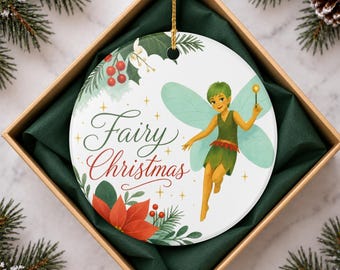 May include: A round white ceramic ornament featuring a green-clad fairy with light blue wings holding a wand. The text Fairy Christmas is written in dark green and red script, surrounded by red berries, green holly leaves, and a red poinsettia.