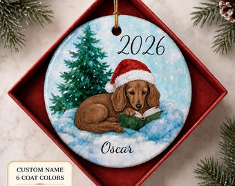 May include: A round ceramic Christmas ornament featuring a brown dachshund wearing a red and white Santa hat, reading a green book in the snow next to a green pine tree. The year 2026 is printed at the top and the name Oscar is printed at the bottom.