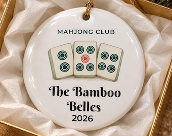 May include: A round white ceramic ornament featuring a gold hanging string. The design includes three mahjong tiles, the text MAHJONG CLUB, The Bamboo Belles, and the year 2026 printed in black and green ink on the front surface.