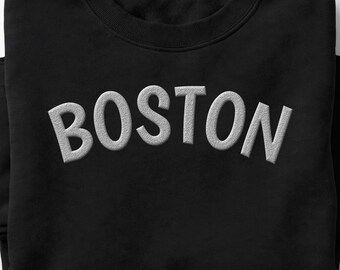 May include: A black crewneck sweatshirt featuring the word BOSTON embroidered in a light gray, textured, arched font across the chest. The garment has a classic round neckline and long sleeves, with the text centered prominently on the front.
