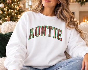 May include: A white long-sleeve crewneck sweatshirt featuring the word AUNTIE printed in an arched, collegiate-style font. The letters are filled with a vertical striped pattern in alternating shades of forest green and bright red.
