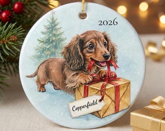 May include: A round white ceramic ornament featuring a long-haired brown dachshund puppy standing next to a gold gift box with a red ribbon. The text 2026 is printed at the top, and a white gift tag with the name Copperfield is attached.