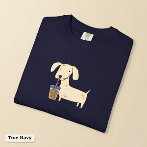 以下が含まれることがあります： ネイビーブルーのTシャツ。青い蓋とピンクのストローが付いた透明なプラスチックカップでアイスコーヒーを飲んでいる、明るいクリーム色のダックスフンドのグラフィックイラストが描かれています。シャツの首元内側には黒い文字が書かれた白いラベルが付いています。