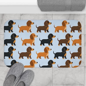 May include: A rectangular bath mat featuring a repeating pattern of cartoon dachshund dogs in various shades of brown and black, set against a light blue background. The dogs are depicted in a standing profile view with simple, clean lines.