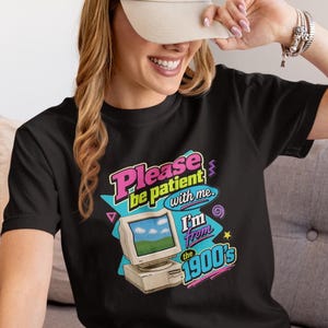 May include: A black t-shirt featuring a graphic of a vintage computer monitor displaying a landscape. The text reads: Please be patient with me. I'm from the 1900's. The design includes colorful accents in pink, yellow, and blue, with geometric shapes.