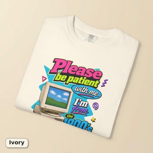 以下が含まれることがあります： アイボリーのTシャツで、青い空と緑の丘が表示されたベージュのヴィンテージコンピューターモニターのグラフィックプリントが施されています。テキストには「Please be patient with me. I'm from the 1900's.」と書かれています。デザインにはピンク、青、紫の幾何学模様が含まれています。