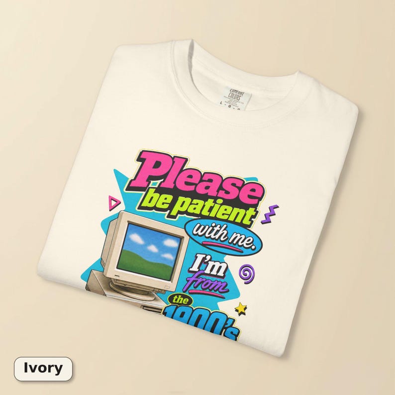 以下が含まれることがあります： アイボリーのTシャツで、青い空と緑の丘が表示されたベージュのヴィンテージコンピューターモニターのグラフィックプリントが施されています。テキストには「Please be patient with me. I'm from the 1900's.」と書かれています。デザインにはピンク、青、紫の幾何学模様が含まれています。