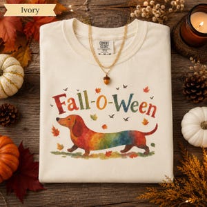 May include: An ivory crewneck t-shirt featuring a colorful watercolor illustration of a dachshund dog and the text Fall-o-Ween. The design includes scattered autumn leaves and small birds. A gold-toned necklace with an acorn pendant rests on the shirt.