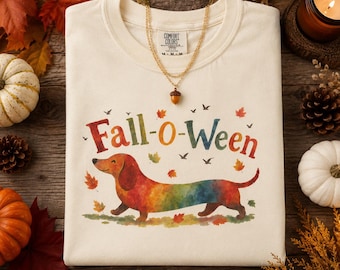 May include: An ivory t-shirt featuring a watercolor-style illustration of a multicolored dachshund dog. The text "Fall-o-Ween" is printed above the dog in a multi-colored font, surrounded by small autumn leaves and birds. A gold necklace is draped over the collar.