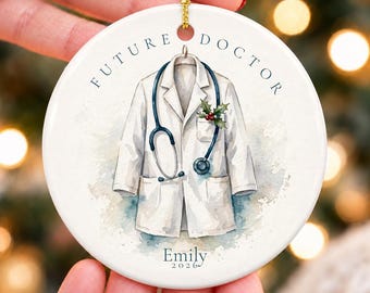 May include: A round white ceramic ornament featuring a watercolor illustration of a white lab coat with a stethoscope draped over it. The text FUTURE DOCTOR is printed above, and the name Emily 2026 is printed below the lab coat.