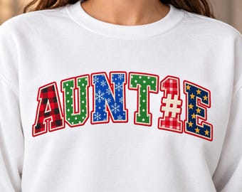May include: A white crewneck sweatshirt featuring the word AUNTIE in large, arched, multi-patterned letters. Each letter has a red outline and a unique design, including red and black buffalo plaid, green polka dots, blue snowflakes, and stars.