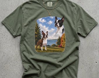 May include: An olive green short-sleeve t-shirt featuring a graphic print of two Boston Terrier dogs in a landscape setting with trees, grass, and a blue sky. A thin gold-tone chain necklace rests on the collar area of the shirt.
