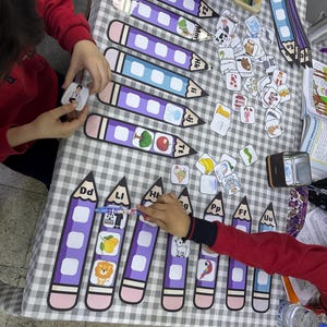 Op de afbeelding: Educatief materiaal voor kinderen, waaronder grote paarse potlooduitsparingen met letters en afbeeldingen, en kleine plaatjeskaarten. De items zijn gerangschikt op een geruite tafelkleed.