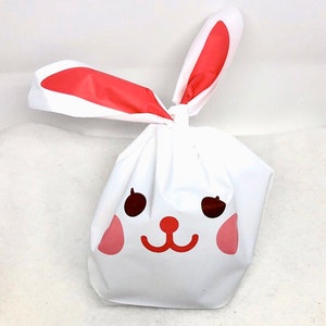 May include: White bunny-shaped gift bag with red ears and a pink and red face. The bag is made of plastic and has a drawstring closure.