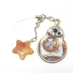 Könnte beinhalten: Ein weißer und orangefarbener Droiden-Charakter-Anhänger mit einem Stern-Anhänger, der an einer silbernen Kette befestigt ist. Die Anhänger sind an einem weißen Handyband befestigt.