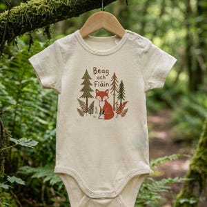 Könnte beinhalten: Ein cremefarbener Kurzarm-Babybody mit einem Grafikdruck von einem Fuchs, Tannenbäumen und Farnen. Der Text "Beag ach Fiáin" ist in Braun über dem Fuchs aufgedruckt. Das Kleidungsstück hat einen Rundhalsausschnitt und zwei Druckknöpfe an der Schulter.