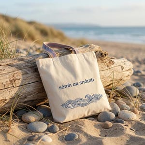Peut inclure: Un sac fourre-tout en toile beige avec les mots "sámh ar snámh" et un motif de vagues bleues. Le sac a une poignée rayée et repose sur un morceau de bois flotté sur une plage de sable. L'océan est visible en arrière-plan.