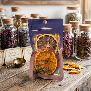 May include: A transparent, iridescent pouch containing dried orange and lemon slices, and green herbs. The pouch is on a wooden surface, with jars of herbs and spices in the background. A sign above reads "Crafted in Small Batches."