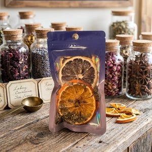 May include: A transparent, iridescent pouch containing dried orange and lemon slices, and green herbs. The pouch is on a wooden surface, with jars of herbs and spices in the background. A sign above reads "Crafted in Small Batches."