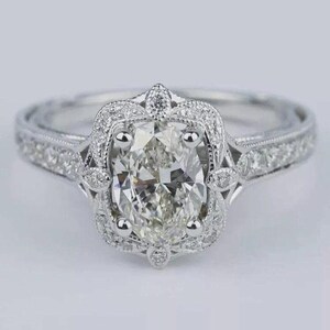 May include: A silver engagement ring with an oval-cut diamond center stone. The diamond is surrounded by a halo of smaller diamonds, and the band is also adorned with diamonds. The ring has an intricate, vintage-inspired design.