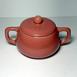 May include: A coral-colored ceramic sugar bowl with a lid and two handles. The lid has a small, round knob. The bowl has a rounded shape and a glossy finish, suitable for kitchen or dining decor.